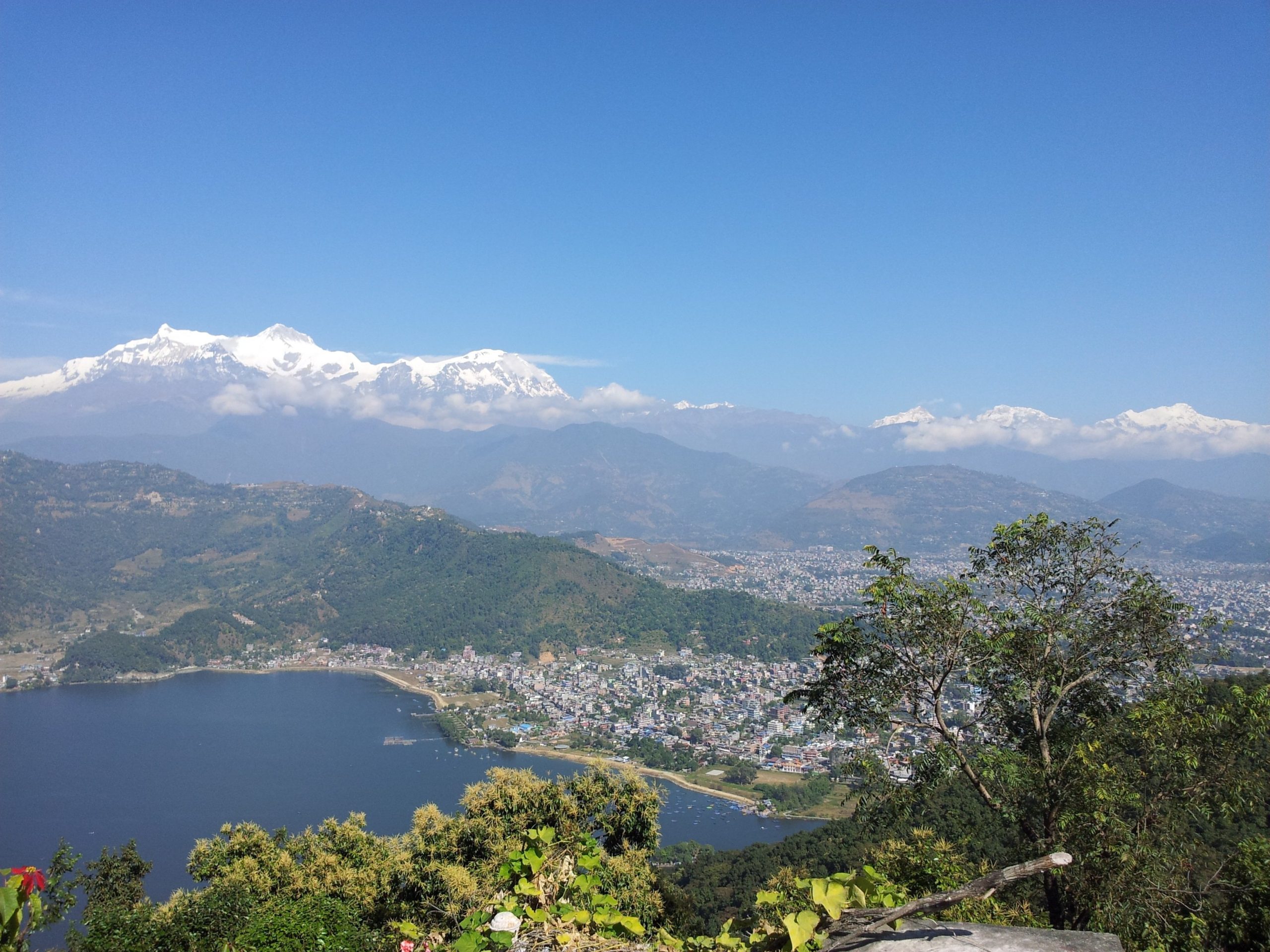 Panoramic view of a lakeside city with a large calm lake in the foreground, green hills surrounding the town, and snow-capped Himalayan mountains under a clear blue sky in the background.