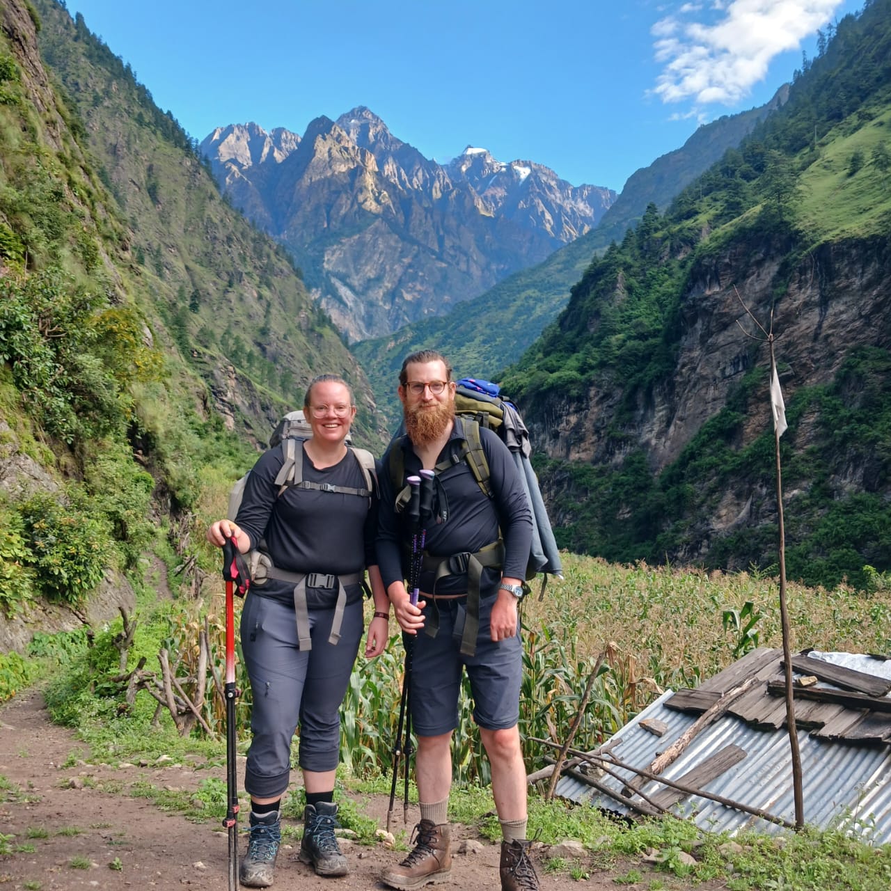 Two trekkers posing with hiking gear in a lush Himalayan valley, with dramatic mountain peaks and blue sky in the background during a trek in Nepal.