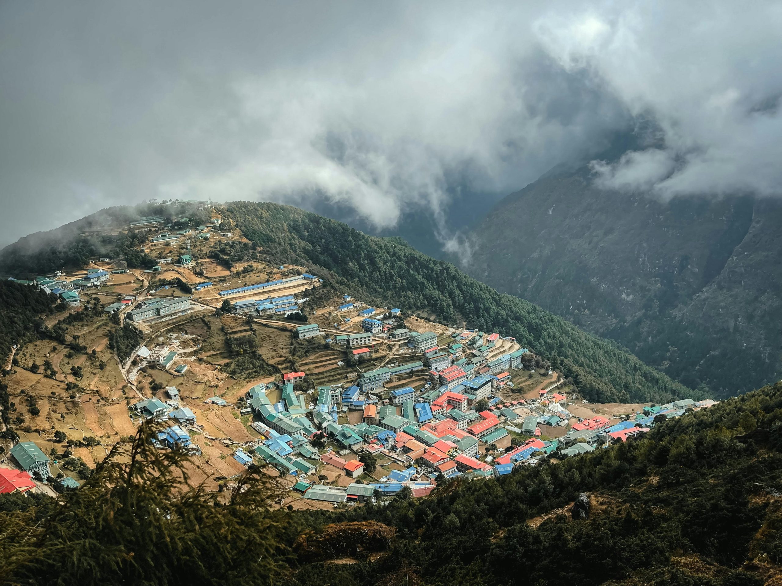A Himalayan village on a steep hillside with colorful rooftops, surrounded by mountains and clouds.