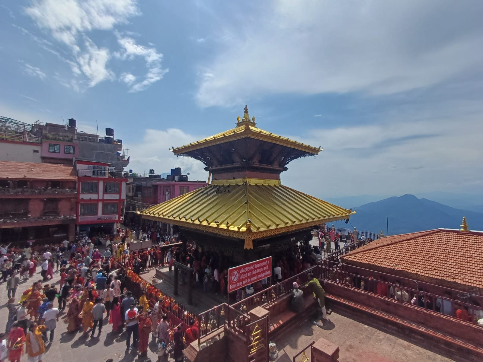 Golden-roofed Buddhist temple surrounded by a busy crowd, colorful prayer offerings, and traditional buildings, with Himalayan mountains and a clear blue sky in the background.