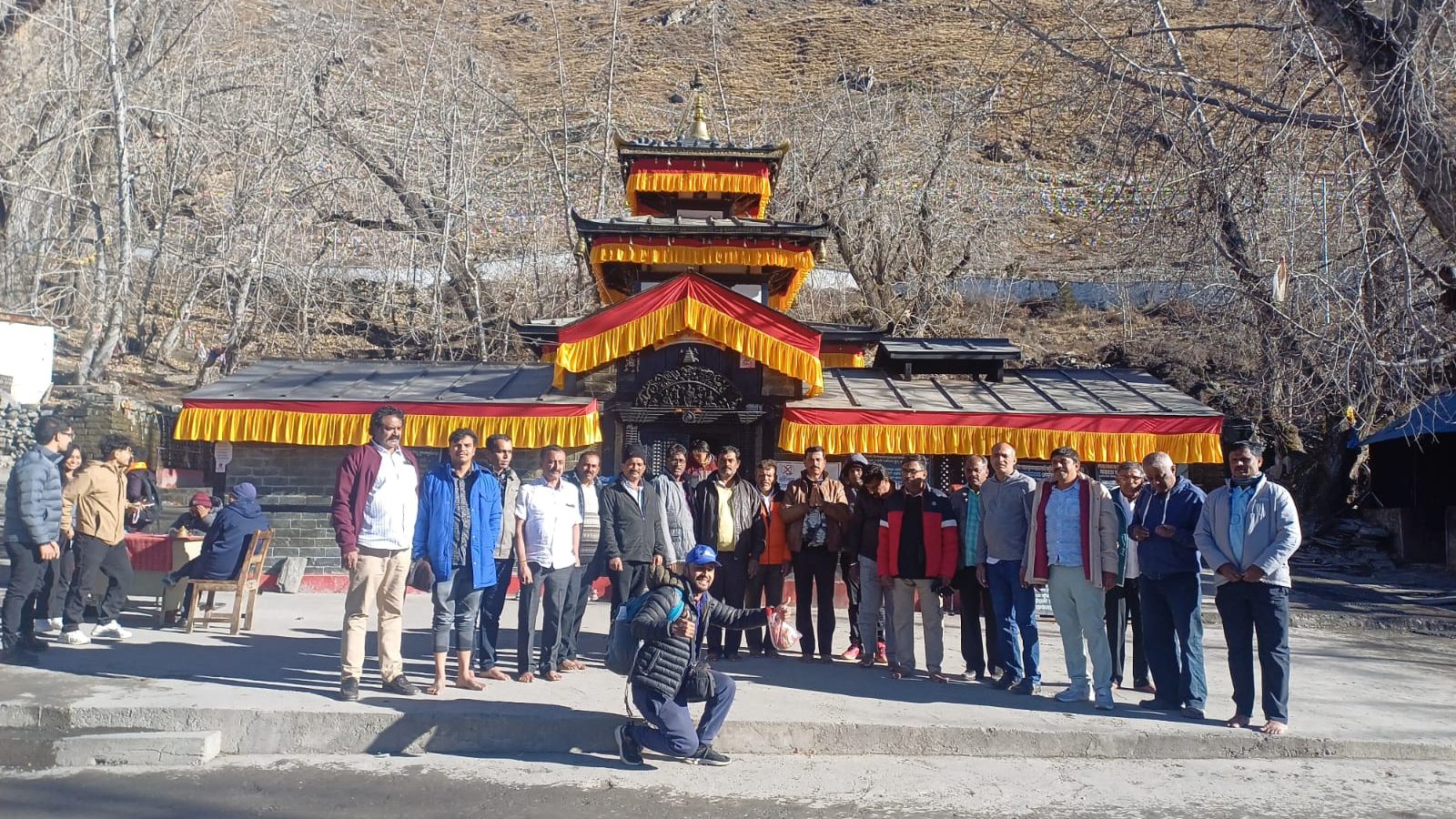 Group of visitors gathered in front of Muktinath Temple, decorated with traditional red and golden fabric, surrounded by leafless trees.